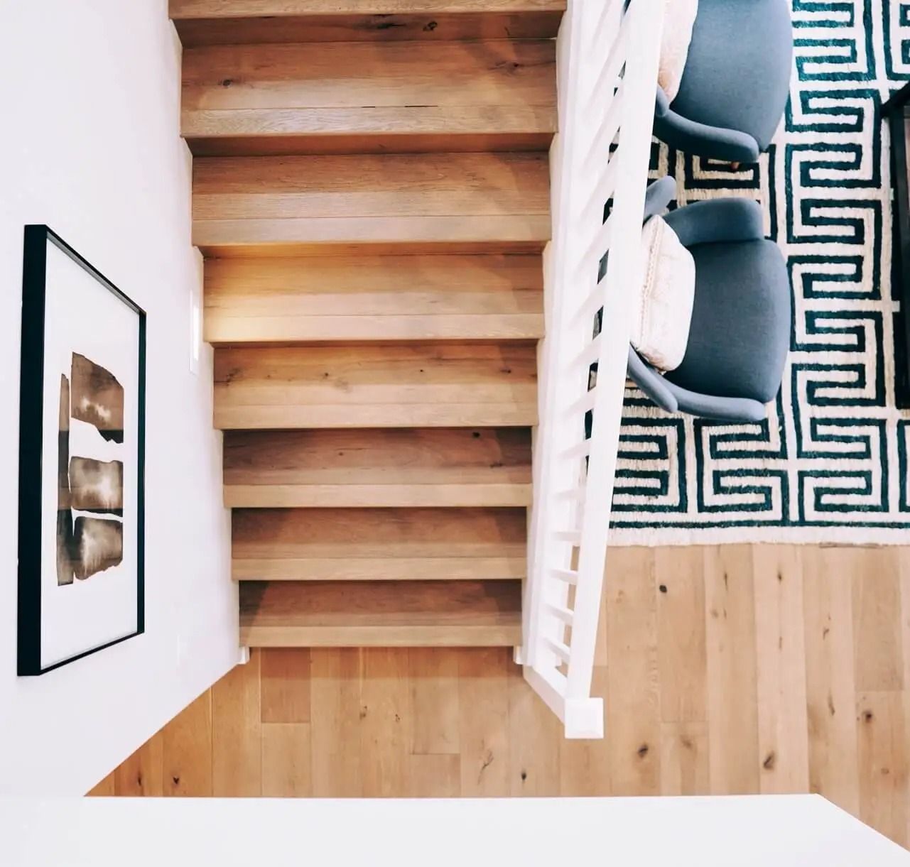 Wooden staircase leads down; white railing and wall; black and white rug; two blue chairs.