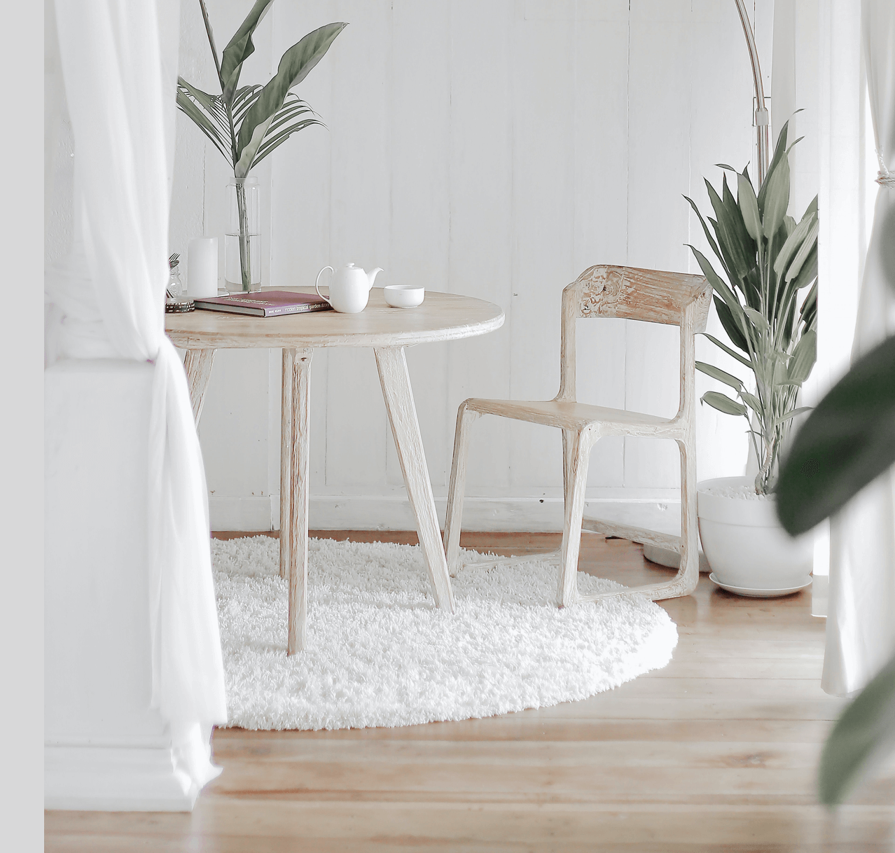White dining area with round table, chair, rug, plants, and sheer curtains.