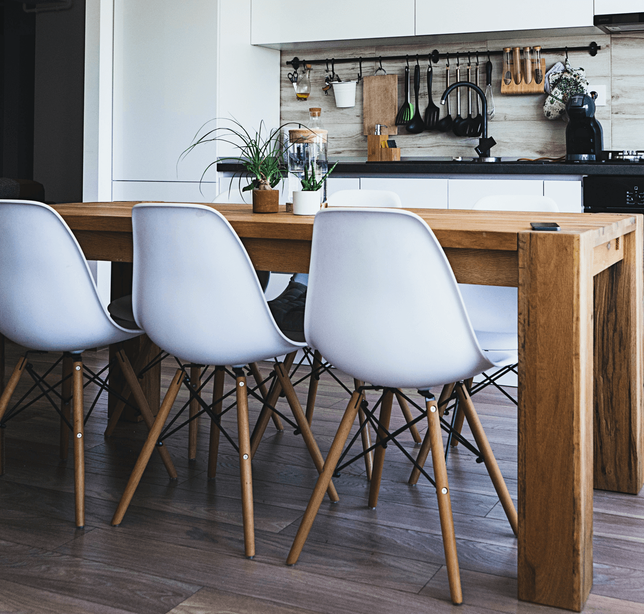 Wooden table with white chairs in a kitchen. Plants on the table, cooking tools on the backsplash.