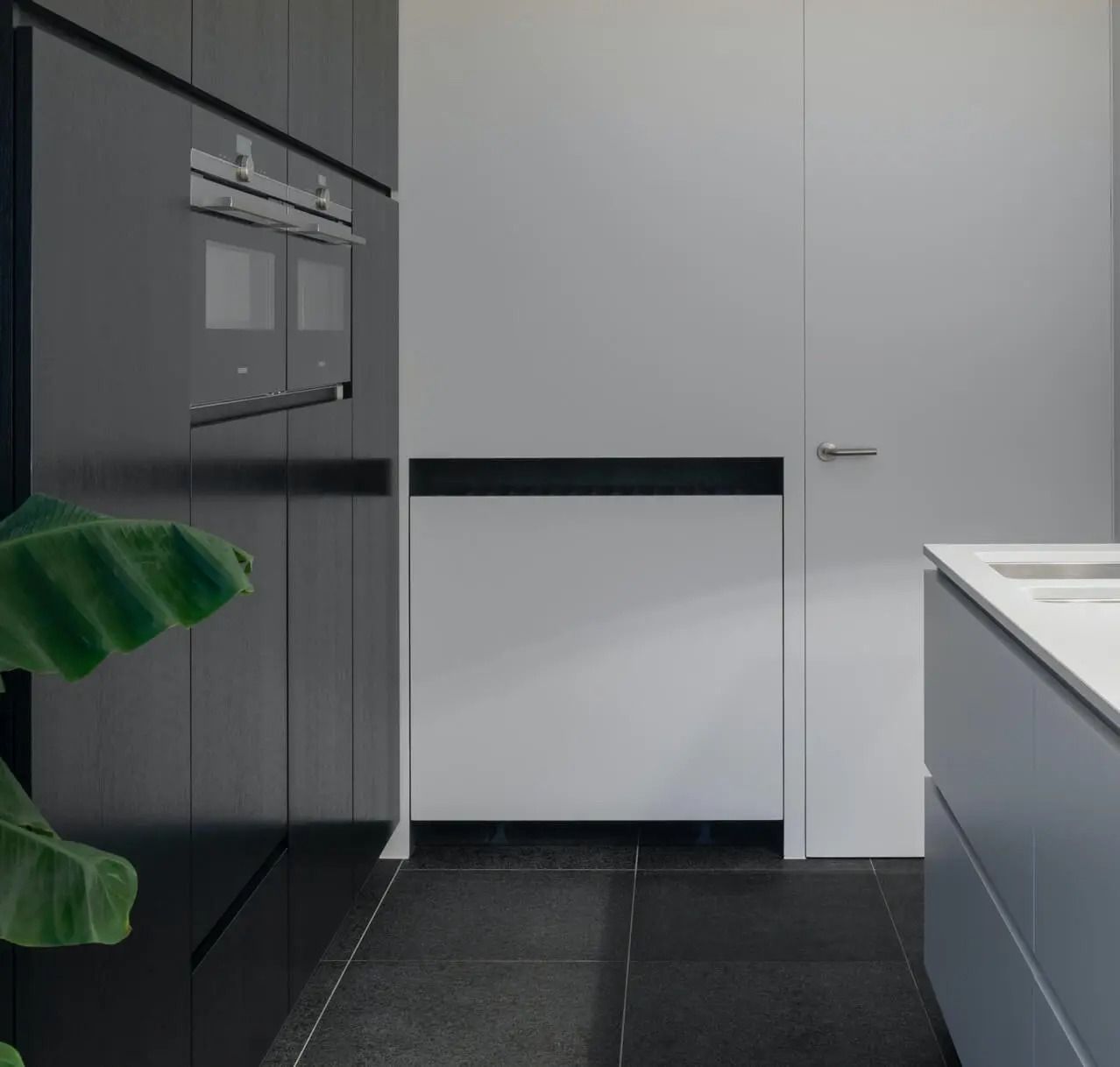 Modern kitchen: dark cabinets, built-in oven, white walls, gray countertop, black tile floor.