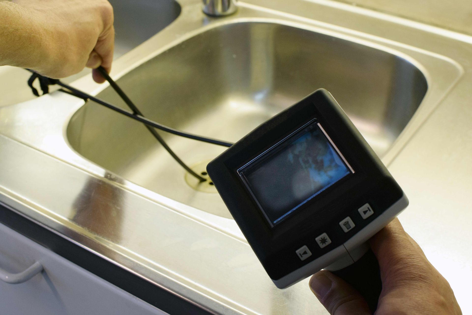 A person using an inspection camera to look inside a sink drain. The camera displays a view on its screen.