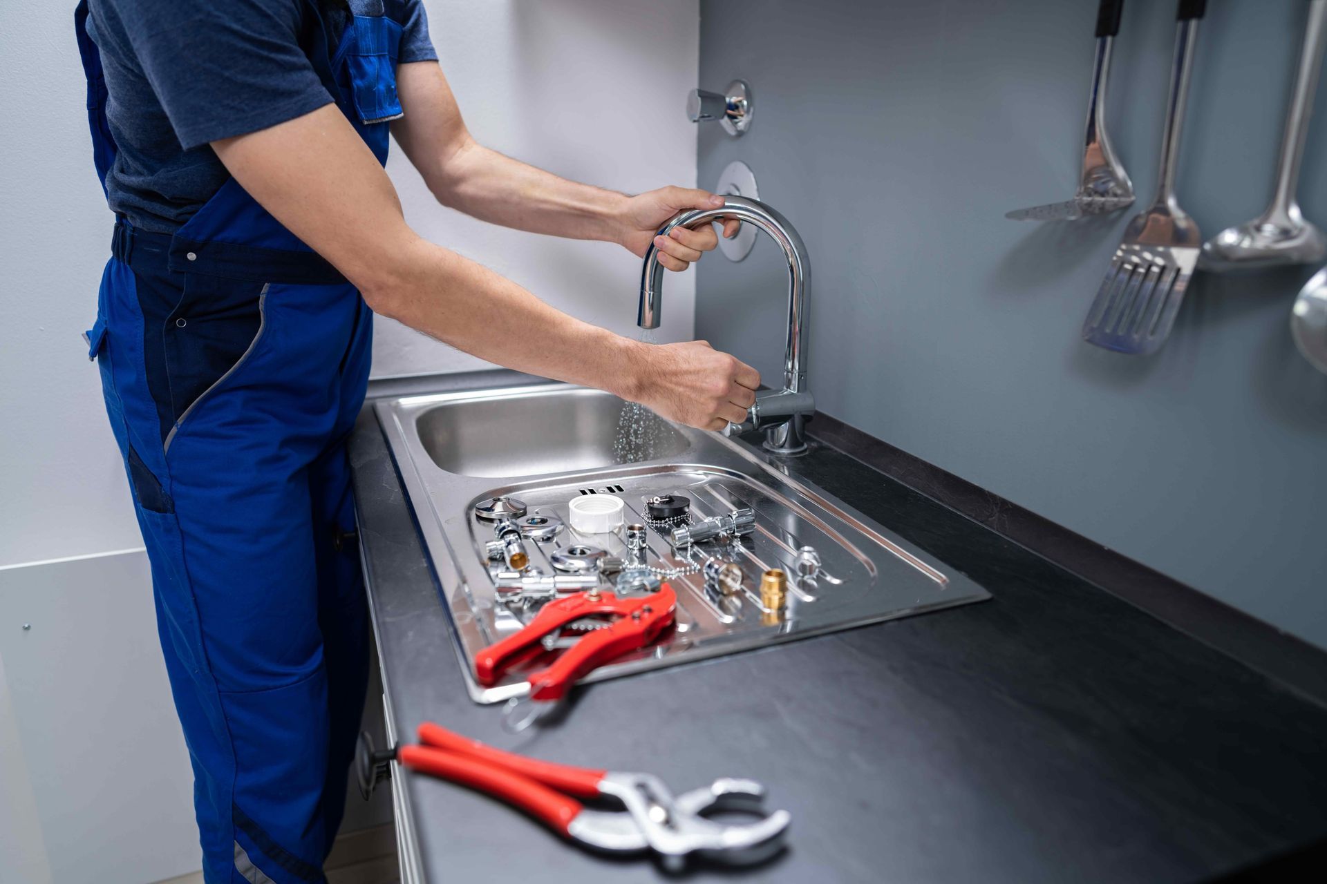 Plumber in blue overalls repairing a kitchen faucet at a stainless steel sink, tools nearby.