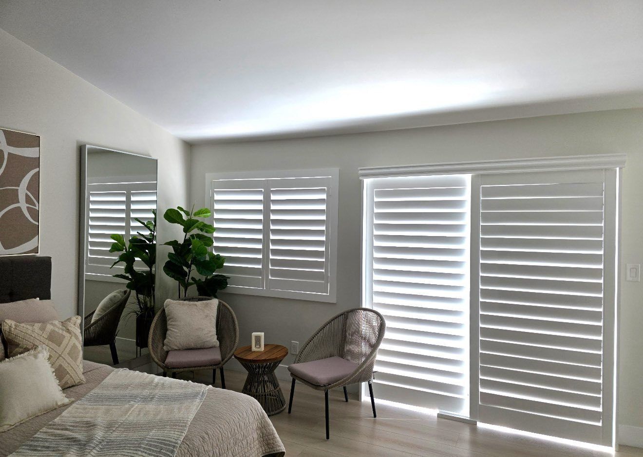 Bedroom with bed, mirror, plant, chairs, and window shutters. Bright, neutral colors.