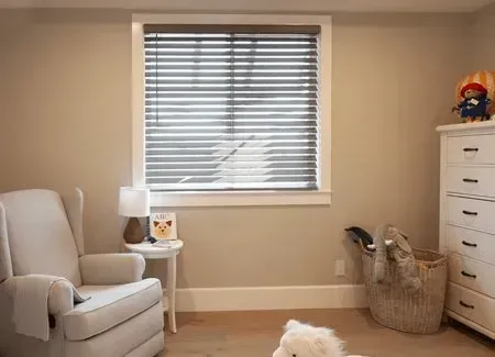 Cozy nursery with a white chair, crib, blinds, and a dresser; beige walls.