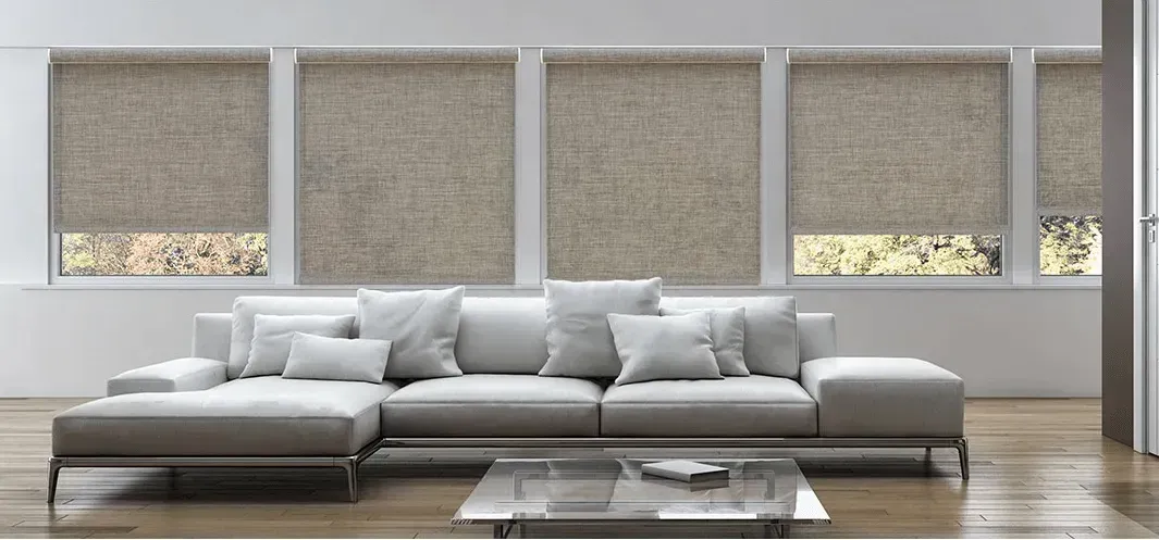 A modern living room with a white sectional sofa, square glass coffee table, and large windows with tan shades.