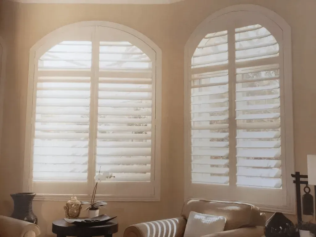 Two arched windows with white shutters in a room with beige walls and furniture.