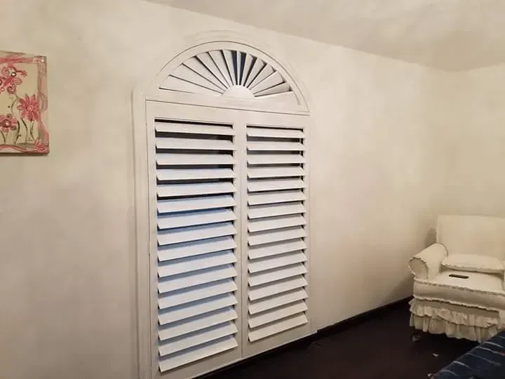 White window shutters with an arched top, partially covering a window on a light-colored wall.