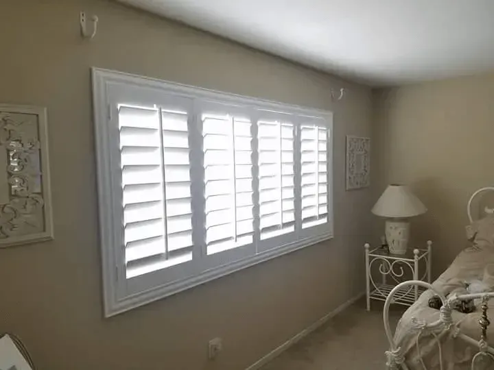 Bedroom window with white shutters, framed by white molding, beige walls, and a nightstand.