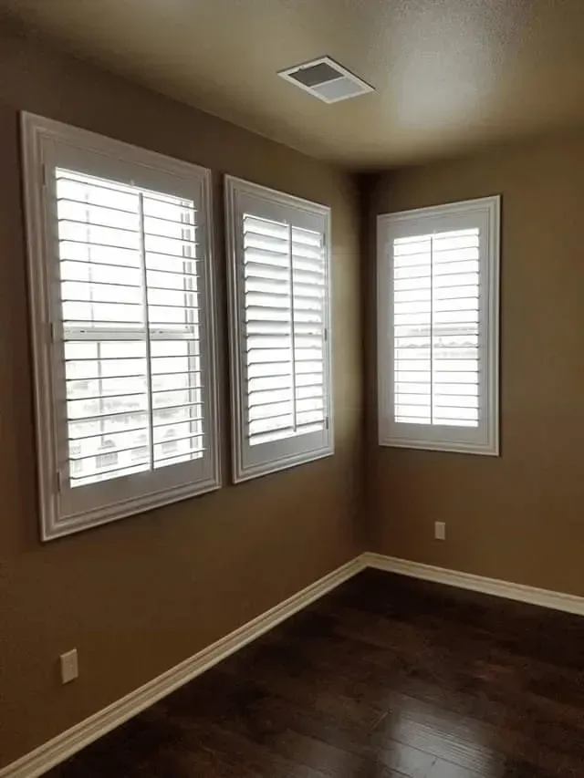 Empty room with three white-framed windows, white shutters, and dark wood floors. Brown walls.
