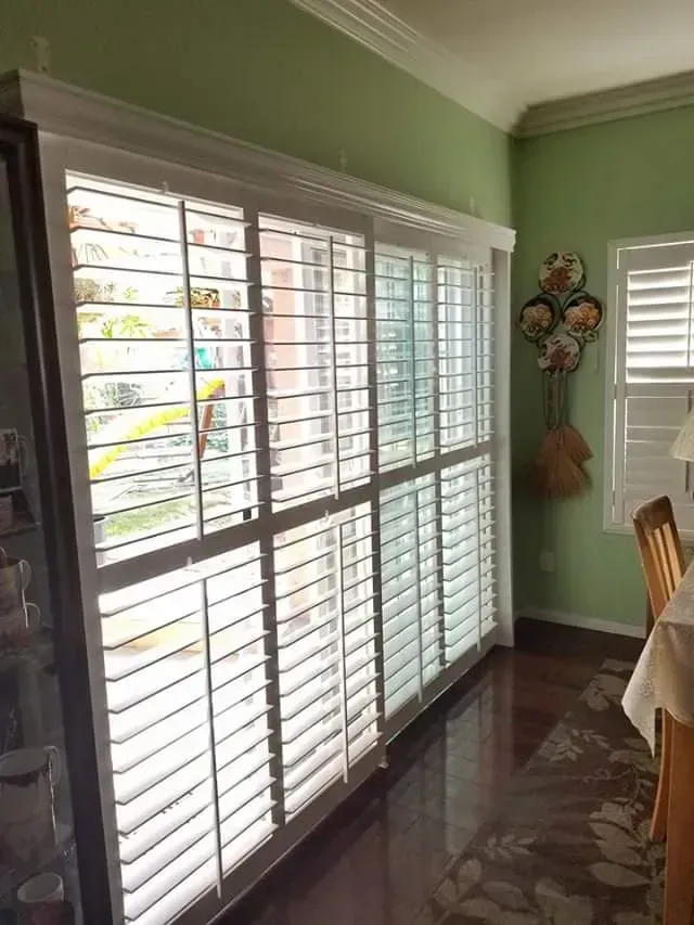 White window shutters partially cover a glass sliding door and a smaller window. Light green walls, dark wood floor.