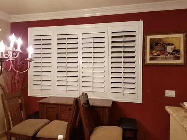 White shutters on a red wall, with a painting and dining chairs in a room.