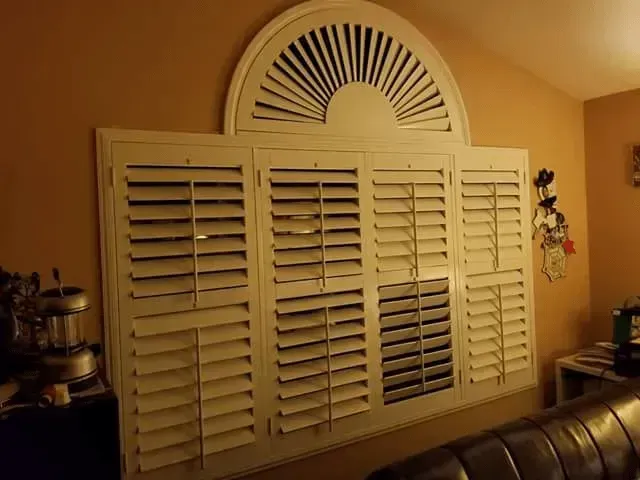 White shutters on a window with arched top, mounted on a tan wall.