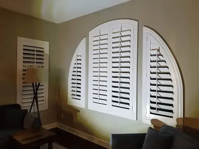 White shutters cover an arched window and a smaller adjacent window, set against a beige wall.