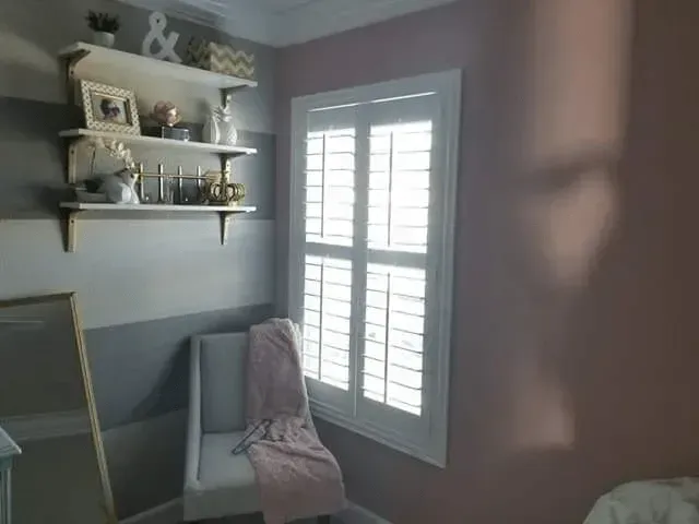 Bedroom corner with shuttered window, gray striped walls, shelves with decor, and a chair with pink blanket.