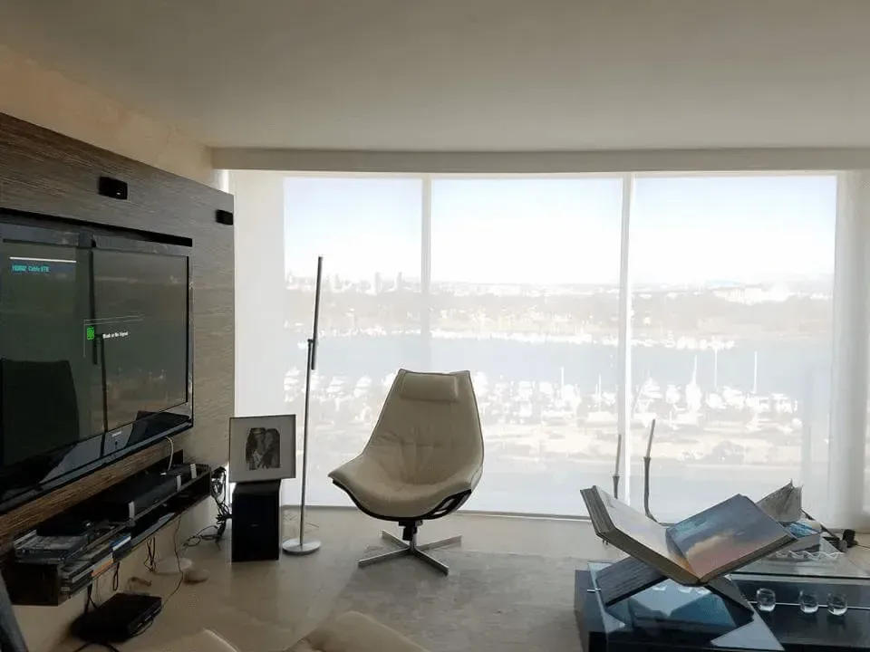 Modern living room with a waterfront view. White chair and table with sheer curtains and a large screen TV.