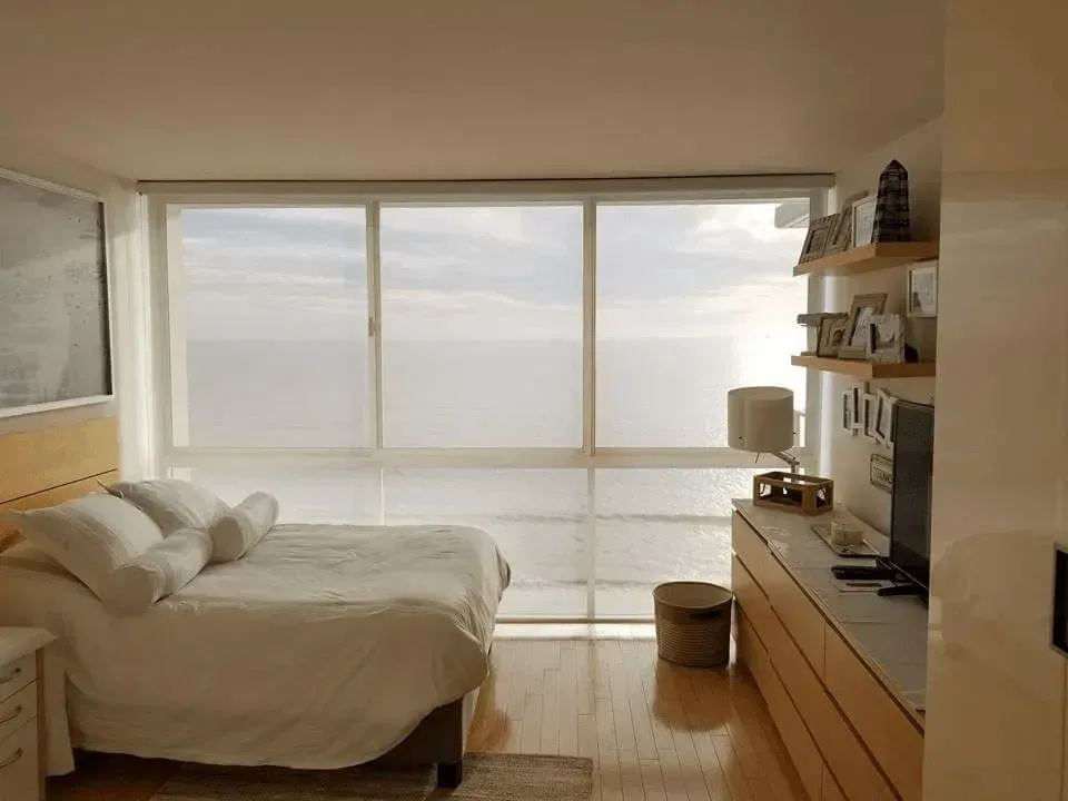 Bedroom with large windows overlooking water, wooden floor, bed with white bedding, and floating shelves.