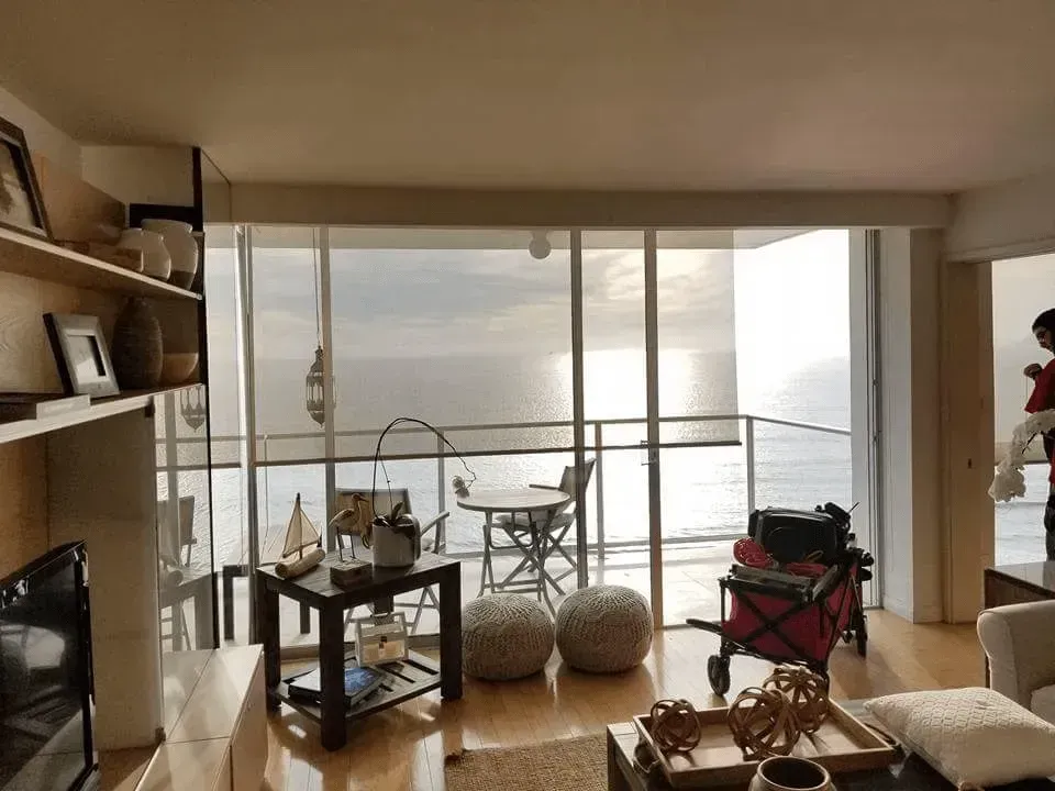 Living room with ocean view, shelves with decor, balcony with table and chairs, sunlight.