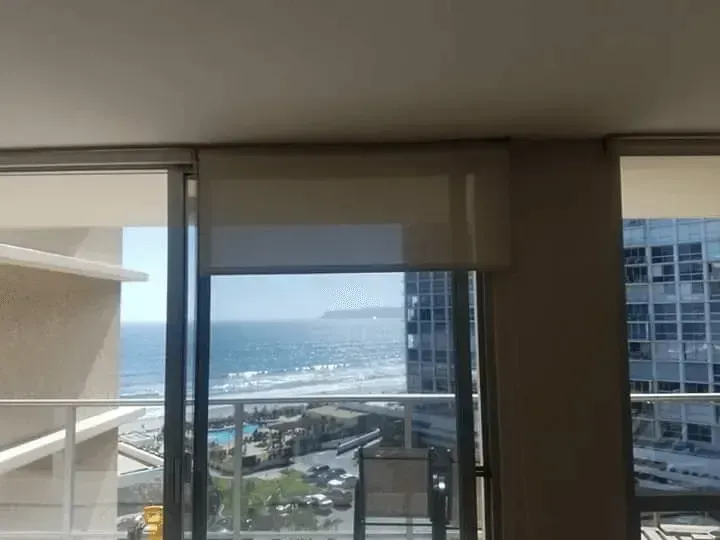 Ocean view from a high-rise window with partially drawn beige roller shade; building and beach in the distance.