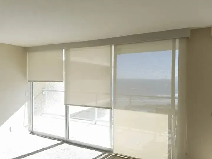 Beige roller shades cover windows; sunlight streams in.