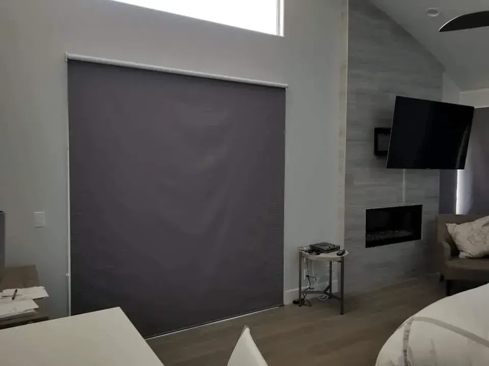 Bedroom with gray roller shade covering a window, TV, fireplace, and bedside table.