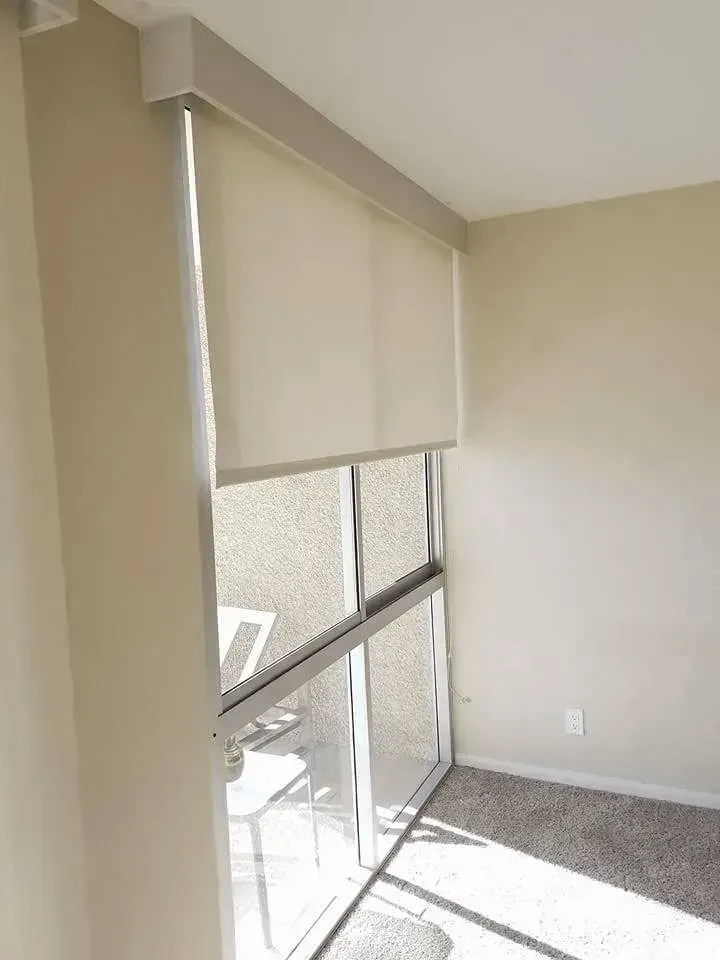 Beige roller shade partially covering a glass sliding door and window with light coming through.