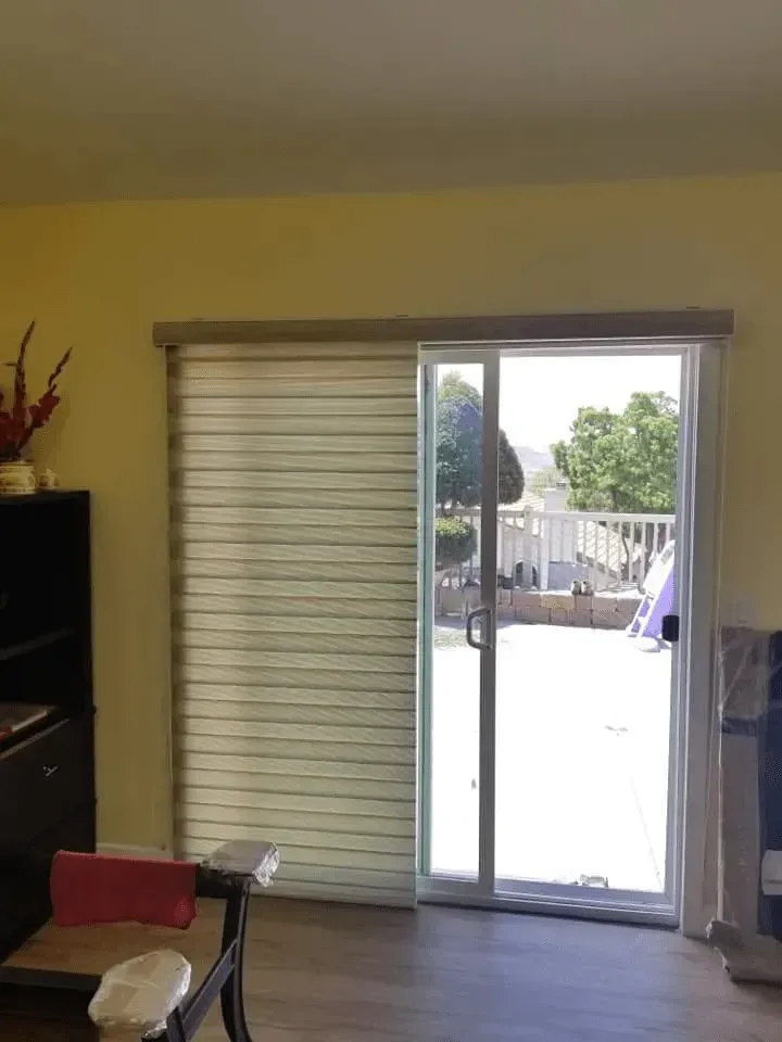 Sliding glass door with partially closed striped shade, opening to a sunny outdoor patio.