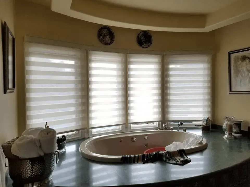 Bathroom with a large oval tub, windows with blinds, and decorative items.