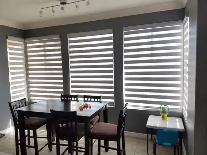 Dining area with a table and chairs in front of three windows with striped blinds; small table to the right.