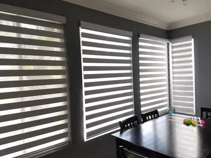 Gray and white zebra blinds covering windows in a room with a dark dining table and chairs.