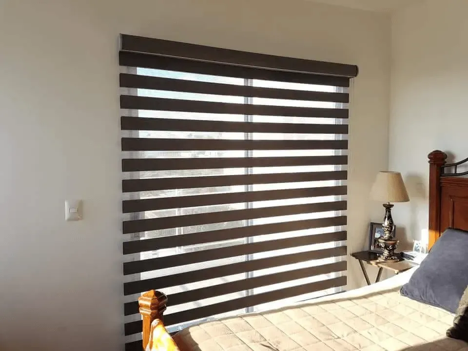 Window with horizontal zebra-striped shades. Bedroom setting with a bed and lamp visible.