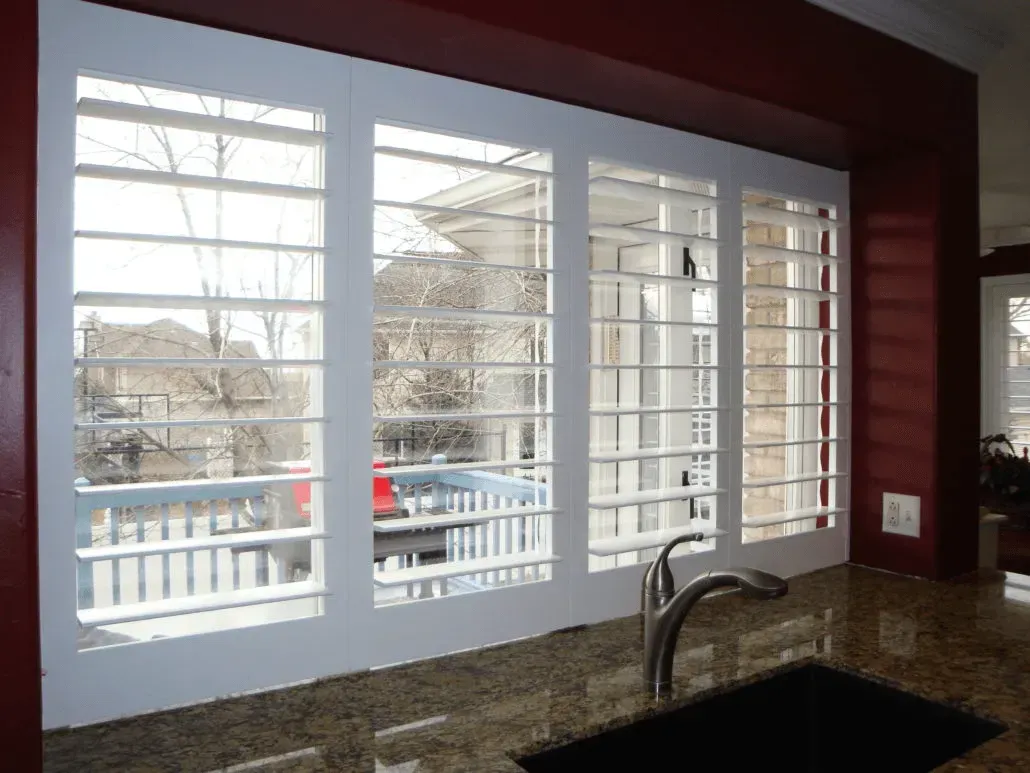 White shutters on kitchen window, showing a balcony and neighborhood in the background. Red wall, dark countertop.