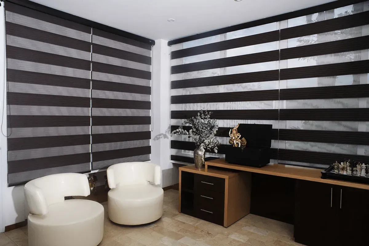 Office interior with layered dark brown and white horizontal blinds. Two white armchairs. Dark wood desk.