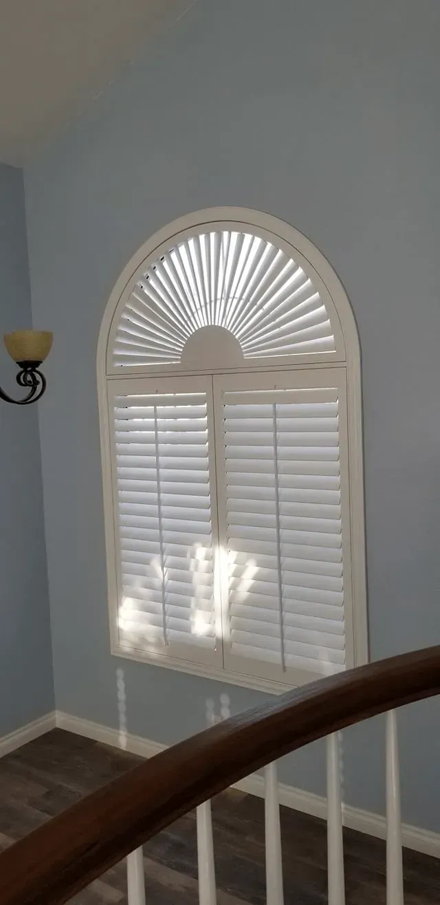 Window with white shutters and a sunburst top, set in blue wall above staircase.