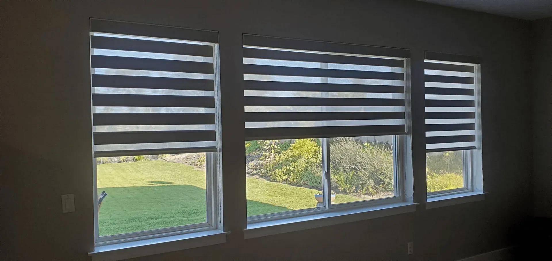 Three windows with zebra-striped blinds partially cover the view of a green lawn and trees.