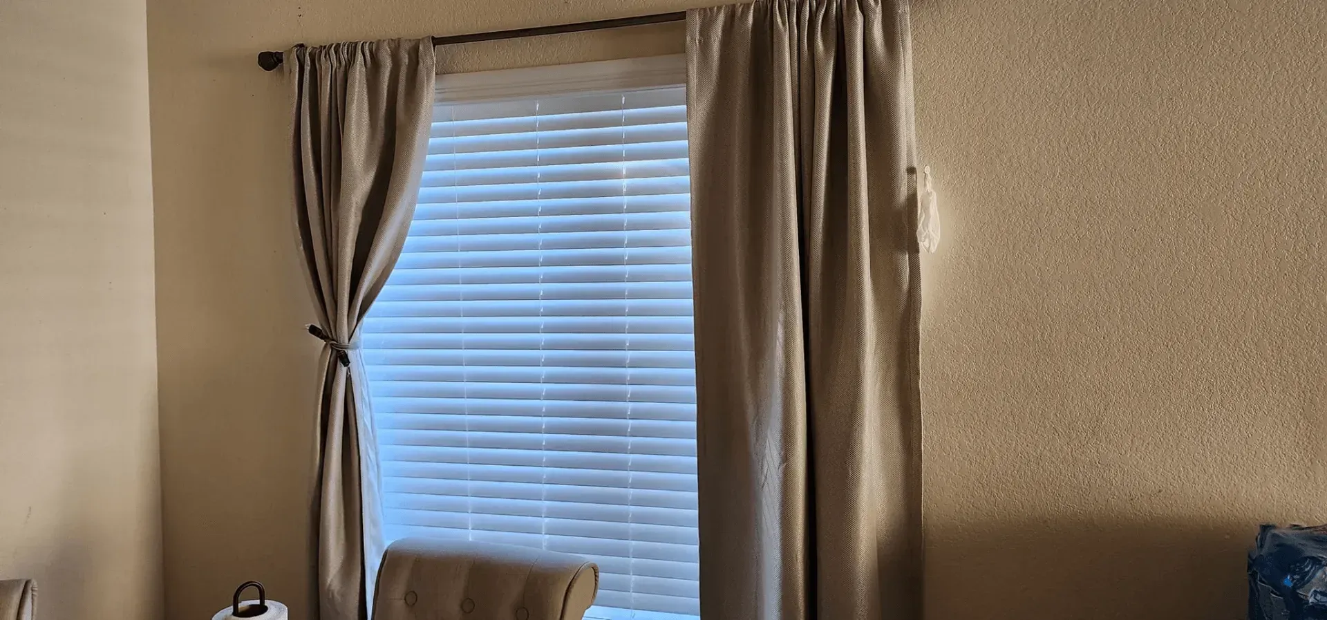 Window with beige curtains and blinds. Beige walls and a chair are also visible.