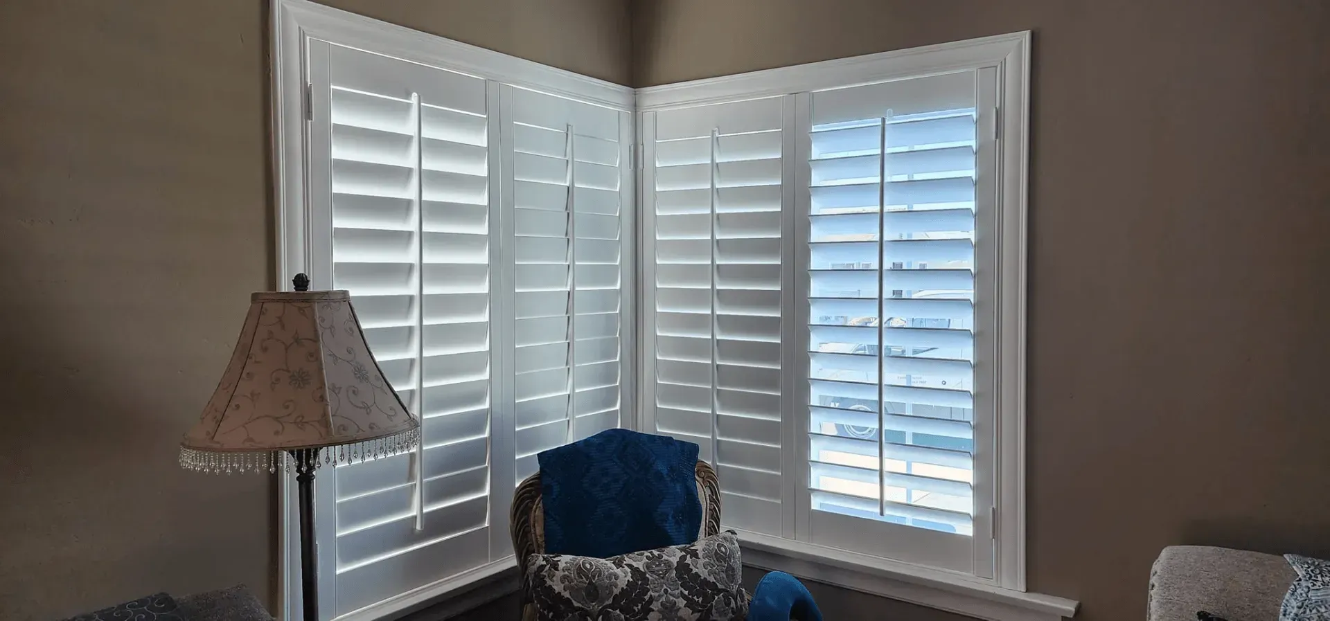 Corner windows with white shutters and a tan lamp with a patterned shade; a blue chair is in the foreground.