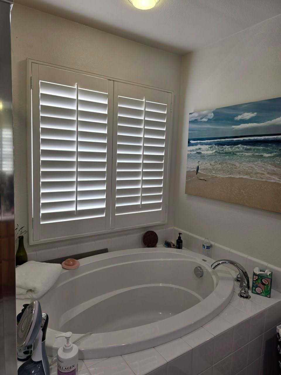 White shuttered windows above a white bathtub in a bathroom, with a beach scene painting.