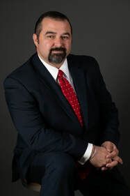 A man in a suit and tie is sitting on a chair with his hands folded.