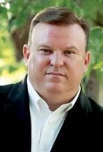 A man in a suit and white shirt is standing in front of a tree.