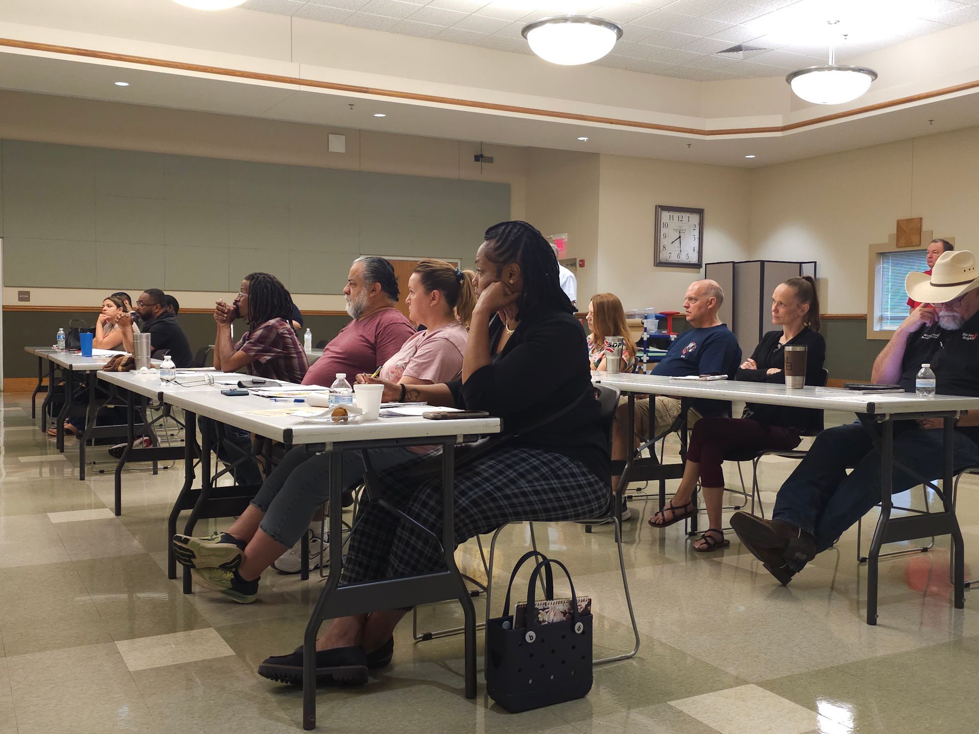 A group of adult people are sitting at tables in a room They are listening and taking notes.