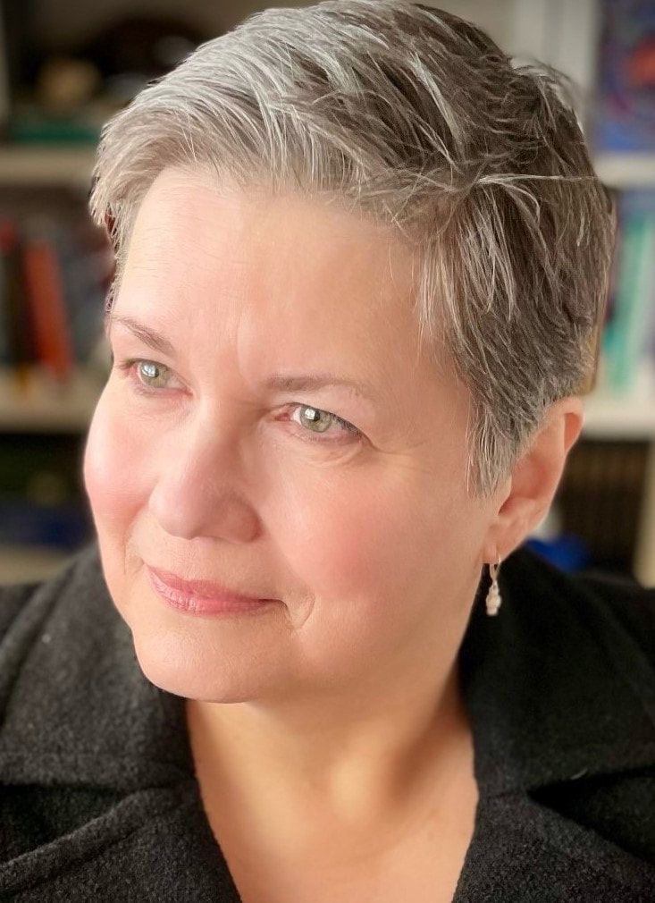 A woman with short gray hair is wearing a black jacket and earrings.