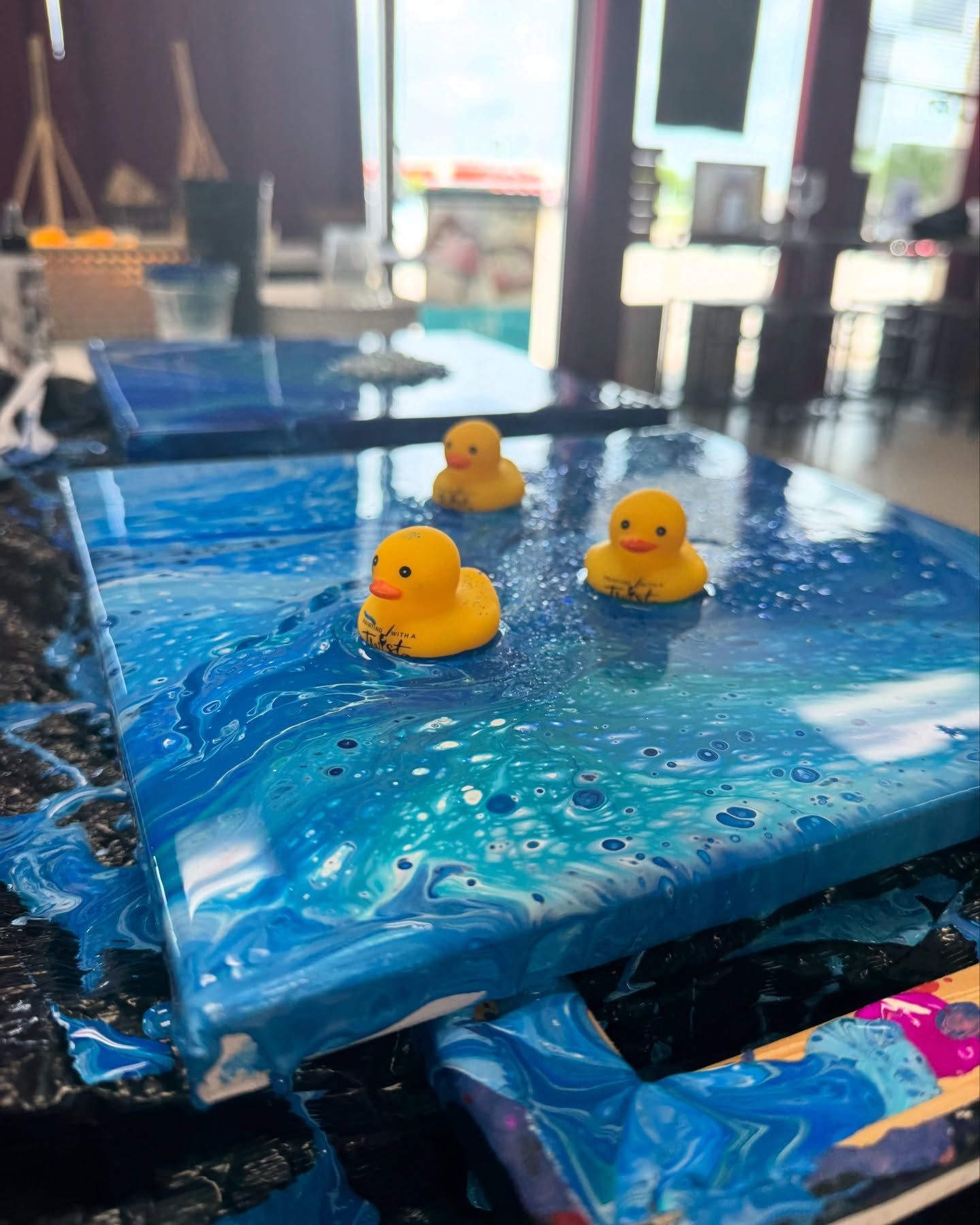 Three yellow rubber ducks are floating on top of a blue pour art painting.