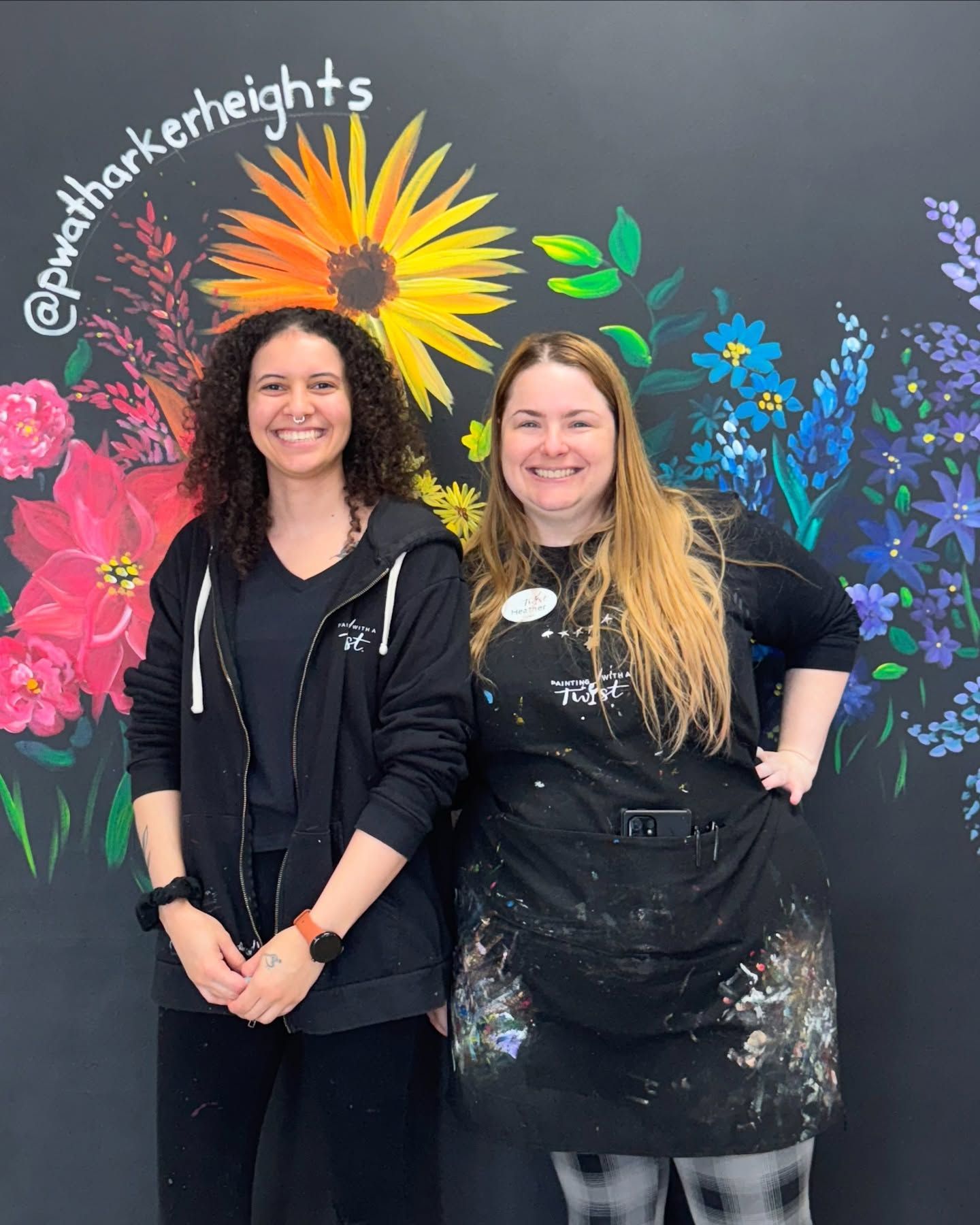 Two women are standing in front of a wall with flowers painted on it.