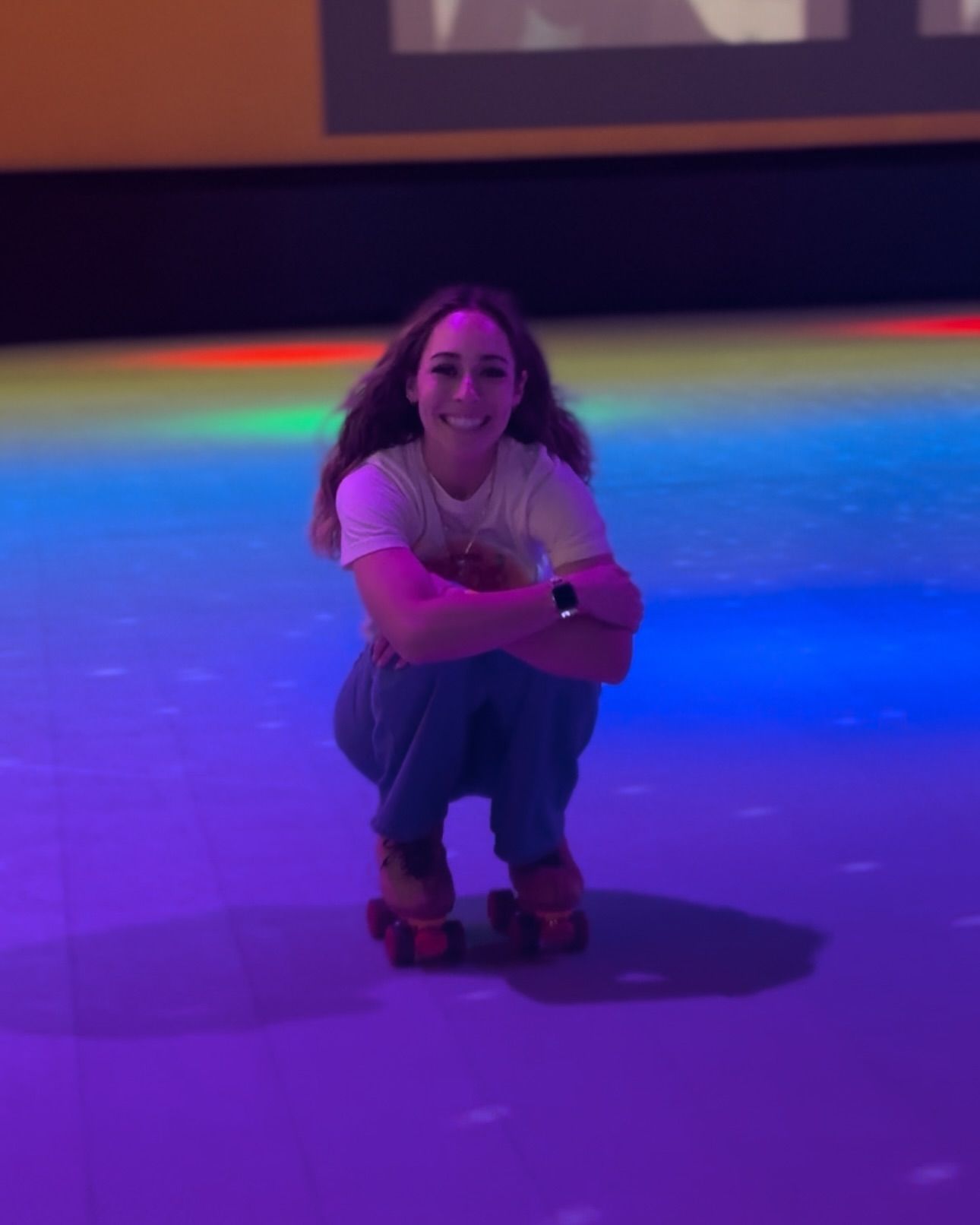 A woman is squatting down on a roller rink wearing roller skates.