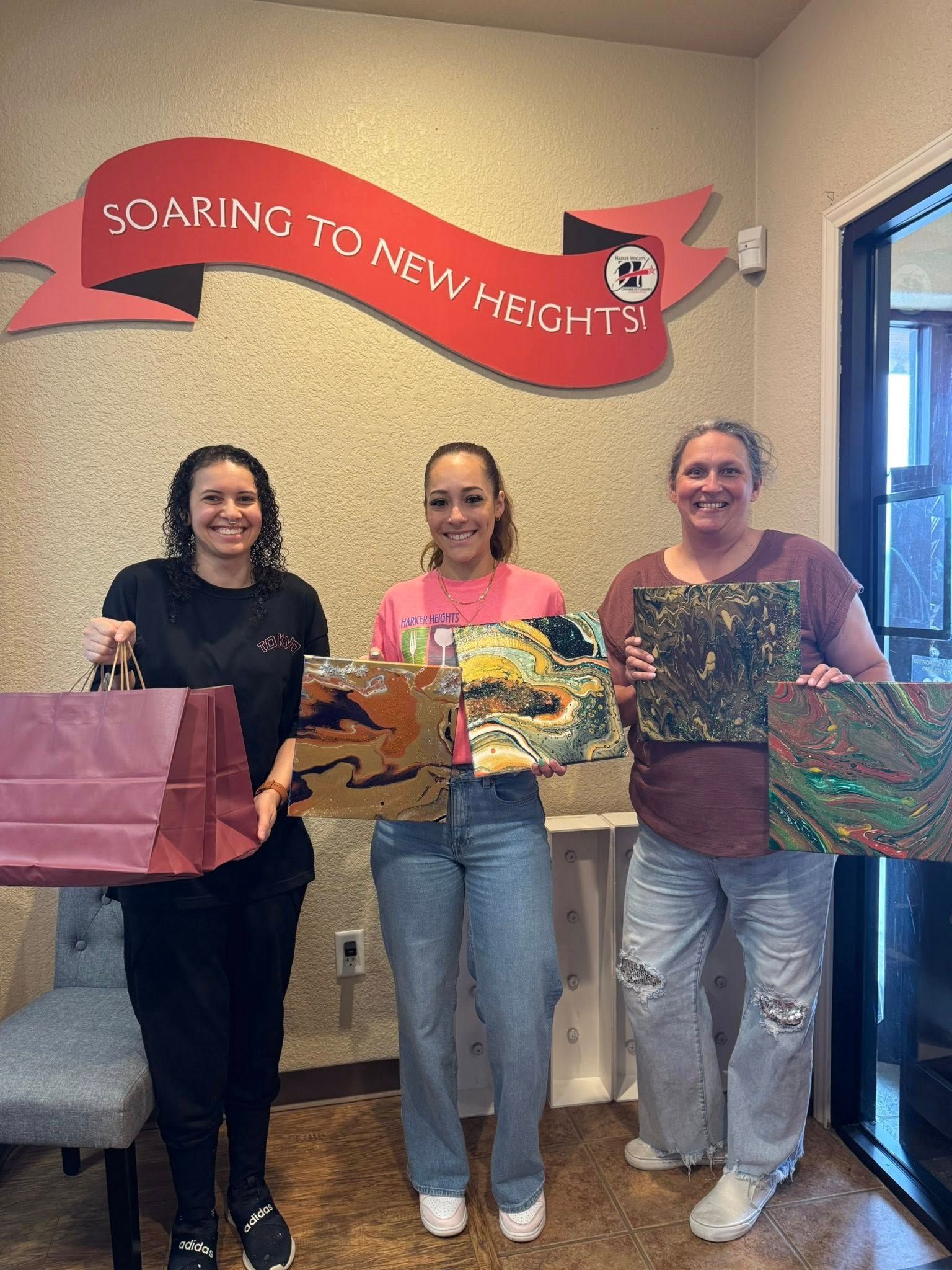 Three women are standing next to each other in a room holding paintings.
