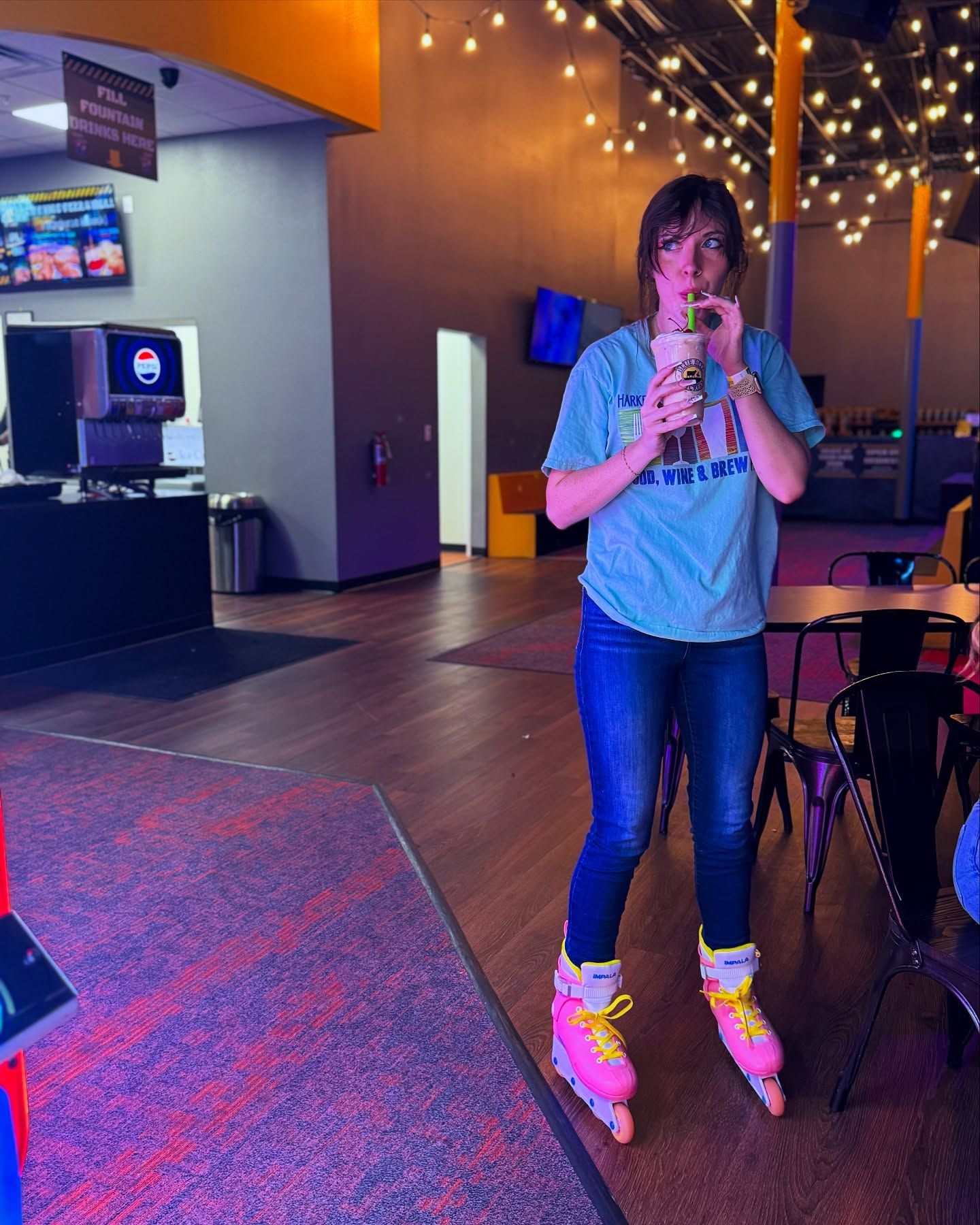 A woman wearing roller skates is standing in a neon lit arcade by the roller rink, sipping a milkshake from a straw.