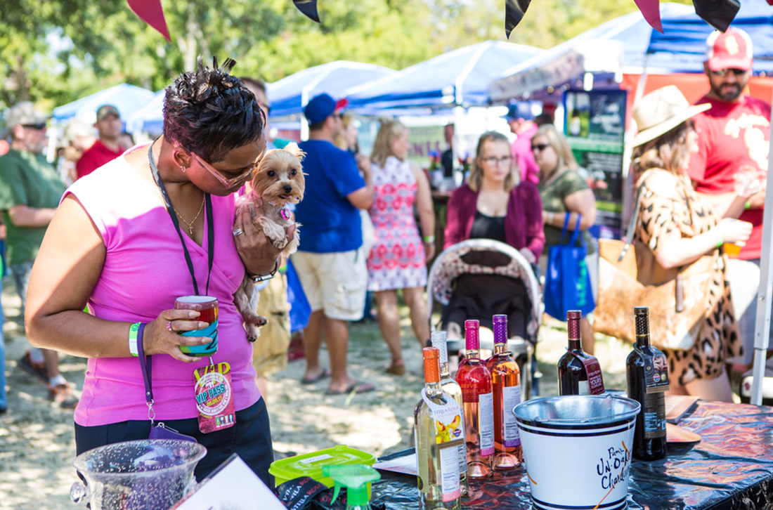 A woman is holding a small dog while standing in front of a table at a festival.