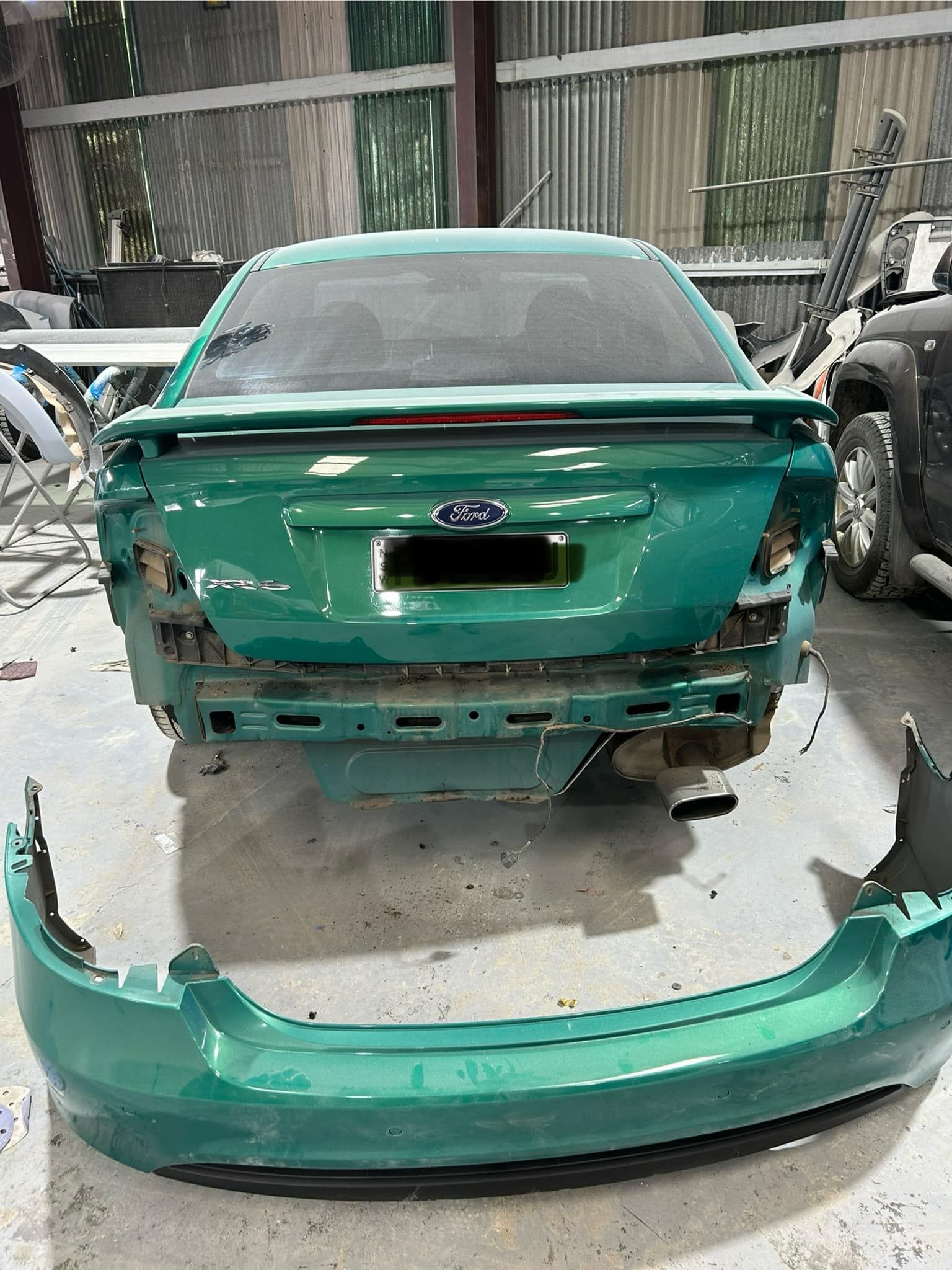 A green car with a bumper missing is sitting in a garage.