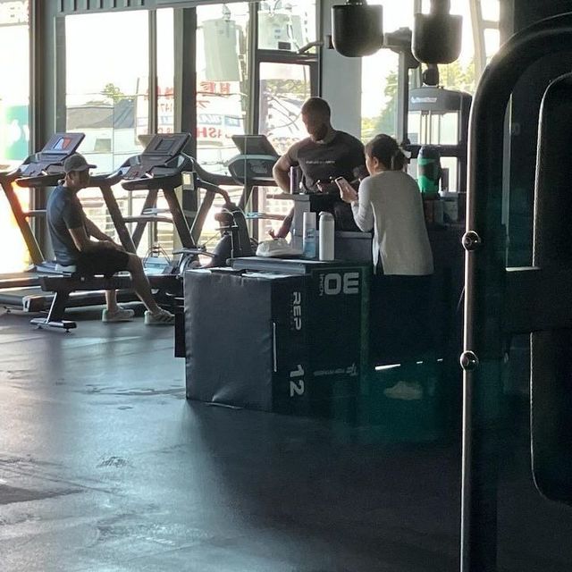 Gym interior: People near equipment. One man on a treadmill, two others near a black box. Bright sunlight.