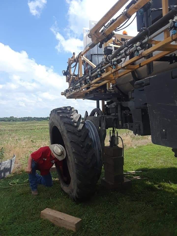 Person in a red shirt and cowboy hat inspecting a large agricultural sprayer tire outdoors under a blue sky.